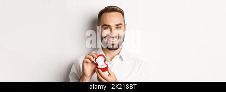 Nahaufnahme des glücklichen, gutaussehenden Mannes, der einen Vorschlag macht, den Ehering in der Box hält und lächelt, ihn bittet, ihn zu heiraten, weißer Hintergrund Stockfoto