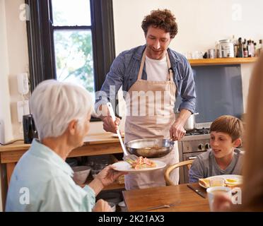 Ein köstliches Frühstück mit freundlicher Genehmigung von Chef Dad. Eine kurze Aufnahme eines glücklichen Mannes, der seiner Familie ein köstlich zubereitetes Frühstück serviert. Stockfoto