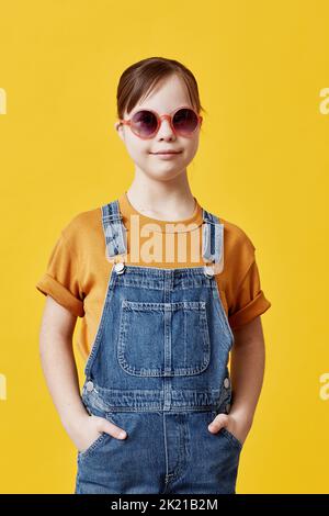 Vertikales Porträt eines niedlichen Mädchens mit Down-Syndrom, das vor gelbem Hintergrund die Kamera anschaut und eine Sonnenbrille trägt Stockfoto