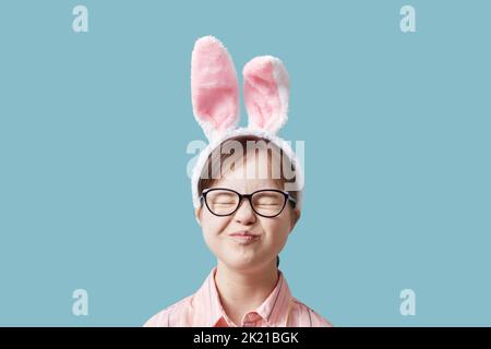 Nettes Mädchen mit Down-Syndrom trägt Osterhasen Ohren vor blauem Hintergrund und macht Gesichter Stockfoto