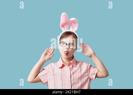 Portrait eines Teenagers mit Down-Syndrom, das Osterhasen-Ohren vor blauem Hintergrund trägt und lustige Gesichter macht Stockfoto