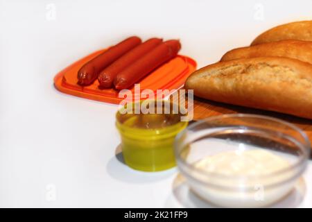 Unschärfe-Zutaten für die Herstellung von hausgemachten Hot Dogs. Würstchen auf orangefarbenem Teller, frisch gebackene Brötchen, Senf und Sauce, serviert auf weißem Hintergrund. Hotdog wi Stockfoto