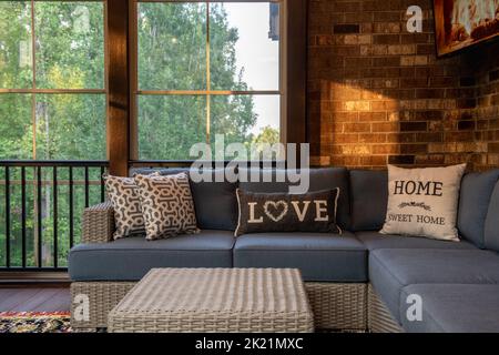 Eine gemütliche Ecke eines Wohnraumes im Freien mit modernen Veranda-Fenstern, Terrassenmöbeln, Kissen, Kissen und Fernseher an Ziegelwänden. Wälder im Hintergrund Stockfoto