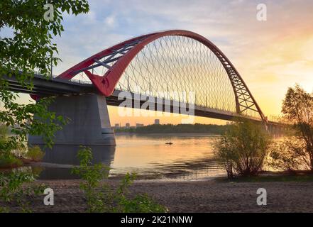 Eine große Bogenbrücke im Morgengrauen. Bugrinsky Brücke über den ob Fluss zwischen den Ästen von Bäumen. Nowosibirsk, Sibirien, Russland, 2022 Stockfoto