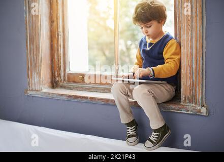 Hes bereits ein Profi mit dem Tablet. Ein kleiner Junge mit einem Tablet, während er auf einer Fensterbank sitzt. Stockfoto