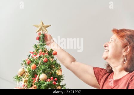 Fröhliche, reife lateinerin, die lächelt, als sie einen goldenen Stern auf ihren Weihnachtsbaum legt, die Freude, die Ferien zu Hause zu verbringen. Stockfoto