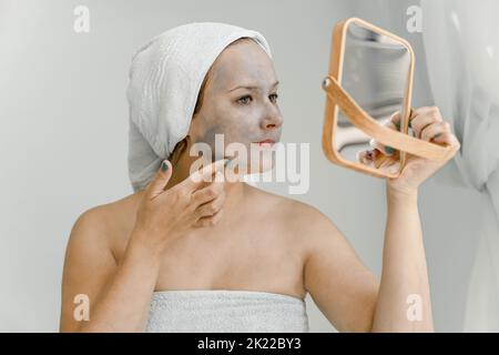 Die junge Frau legt graue kosmetische Tonmaske auf das Gesicht, während sie in Spiegel schaut, ihr Haar und ihren Körper in ein Handtuch gewickelt. Nahaufnahme Stockfoto