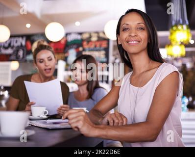 Wer braucht Bürozeiten. Porträt einer jungen Frau, die in einem Restaurant mit zwei ihrer Kollegen im Hintergrund sitzt. Stockfoto