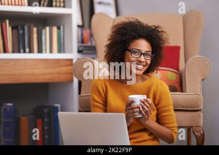 Verbindung wird hergestellt. Porträt einer attraktiven jungen Frau, die Kaffee trinkt, während sie an ihrem Laptop arbeitet. Stockfoto