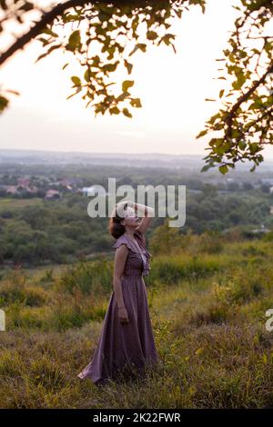 Schöne junge lächelnde Mädchen in einem langen braunen Kleid steht entlang des Rasens. Glückliche Frau geht bei Sonnenuntergang auf einem Hügel mit Blick auf den Fluss. Konzept des havi Stockfoto