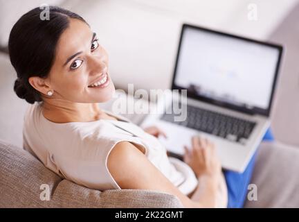 Immer und überall online. Über die Schulter einer jungen Frau, die mit einem Laptop auf einem Sofa sitzt. Stockfoto