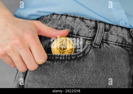 Ethereum Goldmünze in weiblicher Nahaufnahme. Eine Frauenhand steckt ein goldenes ethereum in eine Jeanstasche. Kryptowährung - Ukraine, Izmail, 03.24.2022 Stockfoto