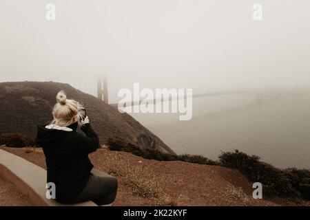 Frau und Tourist fotografieren mit dem Mobiltelefon die Golden Gate Bridge und die Landschaft der San Francisco Bay an einem nebligen und bewölkten Tag. Stockfoto