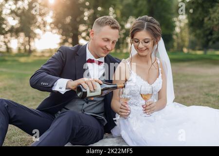 Portrait eines Ehepaares, das im Sommer auf Gras im Park sitzt. Junger Mann Bräutigam Gießen Wein aus der Flasche in Gläser. Stockfoto
