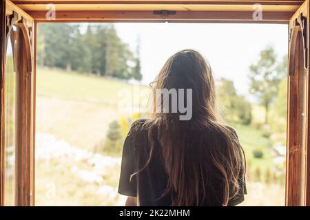 Rückansicht der langhaarigen Brünette Frau, die aus dem Fenster schaut und die Landschaft mit ländlichen Blick mit Bäumen und Hügeln betrachtet. Stockfoto