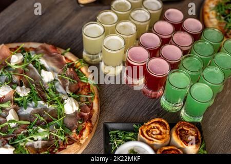 Pizza mit Mozzarella-Käse, Speck, Fleisch auf dem Kaffeetisch. Alkohol bunte rote, gelbe und grüne Gelee Getränke und Brötchen Stockfoto