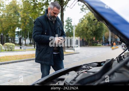 Ein älterer Mann neben der erhöhten Haube eines kaputten Autos ruft auf einem Mobiltelefon um technischen Support Stockfoto