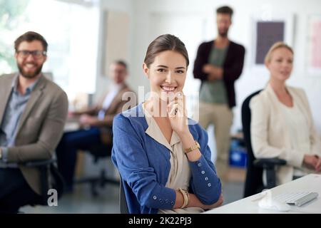 Geschätzter Teil eines kreativen Teams. Junge Geschäftsfrau lächelt mit Kollegen im Hintergrund an der Kamera. Stockfoto