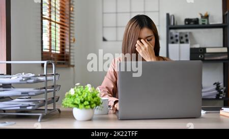 Gestresste, tausendjährige asiatische Geschäftsfrau oder Chefin, die an ihrem Schreibtisch an ihrer Geschäftsaufgabe arbeitet und unter Kopfschmerzen oder Augenschmerzen leidet. Stockfoto