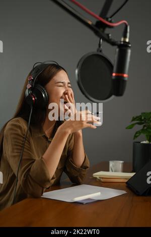 Fröhliche junge Radiomoderatorin lacht, während sie Gäste interviewt und im Heimstudio Live-Podcasts aufgenommen hat Stockfoto