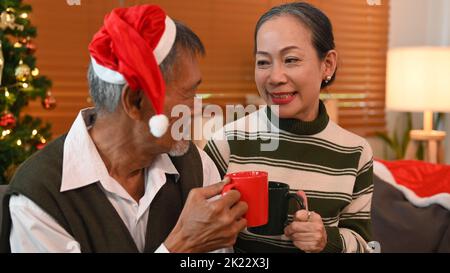 Liebevolles Seniorenpaar, das Spaß hat, heiße Schokolade zu trinken, während es am geschmückten Weihnachtsbaum im Wohnzimmer sitzt Stockfoto