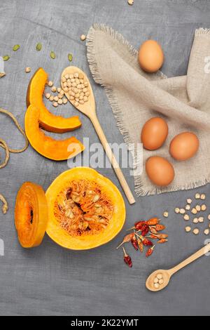 Eier auf Serviette. Kürbisstücke in Scheiben und halber Kürbis. Kichererbsen in Holzlöffel. Flach liegend. Grauer Hintergrund. Stockfoto