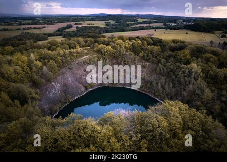 Kleiner blauer Krater-See eines ruhenden Vulkans, umgeben von Wald in Herbstfarben und Bergen am Horizont, in Deutschland Stockfoto