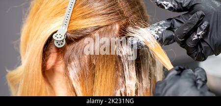 Die Hände des Friseurs tragen den Farbstoff mit Handschuhen auf das rote Haar einer jungen rothaarigen Frau in einem Friseursalon aus nächster Nähe auf Stockfoto