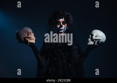 Vorderansicht der Frau in halloween Make-up und schwarzes Kostüm stehen mit Totenköpfen auf dunkelblauem Hintergrund, Stockbild Stockfoto