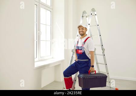 Glücklicher männlicher Arbeiter in Uniform, der im leeren Raum steht, Werkzeugkasten hält und lächelt Stockfoto