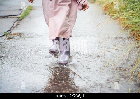 Kleines Mädchen in rosa wasserdichter Regenjacke, lila Gummistiefel lustige Sprünge durch Pfützen auf der Straße bei regnerischem Tageswetter. Frühling, Herbst. Kinder Stockfoto