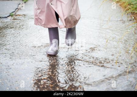 Kleines Mädchen in rosa wasserdichter Regenjacke, lila Gummistiefel lustige Sprünge durch Pfützen auf der Straße bei regnerischem Tageswetter. Frühling, Herbst. Kinder Stockfoto