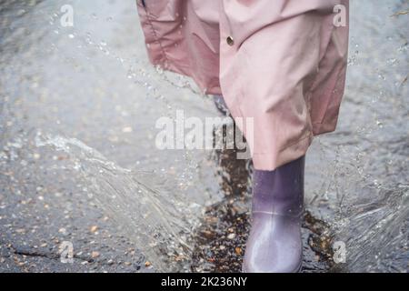 Kleines Mädchen in rosa wasserdichter Regenjacke, lila Gummistiefel lustige Sprünge durch Pfützen auf der Straße bei regnerischem Tageswetter. Frühling, Herbst. Kinder Stockfoto