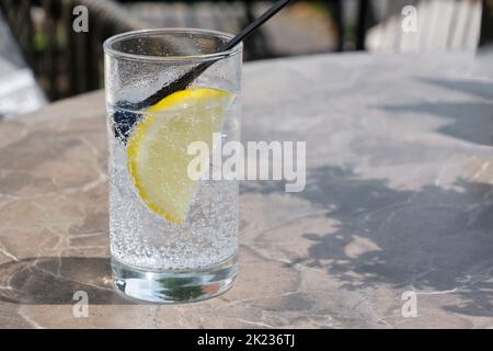 Ein Glas kalter, funkelnder Limonade mit einer Scheibe gelber Zitrone auf einem Tisch in der Sonne. Bild mit selektivem Fokus. Stockfoto