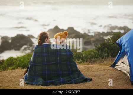 Es wird gemütlich. Rückansicht eines anhänglichen jungen Paares, das in eine Decke gehüllt wurde, auf ihrem Campingplatz. Stockfoto