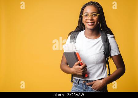 Inspirierende multirassische Studentin in weißem T-Shirt und Jeans mit Rucksack isoliert auf gelb, Ausländer Frau in Brille stolz, Universitätsmitglied zu sein, Blick auf die Kamera im Innenbereich stehen Stockfoto