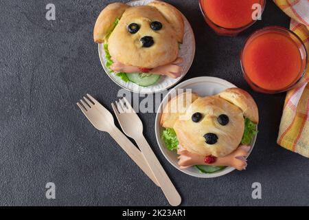 Fun Food für Kinder - zwei Sandwiches in Form eines Hundes mit Gurken, Würstchen und Tomatensaft auf grauem Hintergrund, Draufsicht Stockfoto