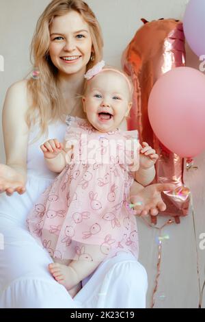 Vertikale lachende und täuschende blonde Frau, Mutter hält Baby in rosa Kleid und Blume Bandelett, feiert ein Jahr Stockfoto