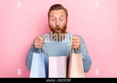 Foto von positiven schönen Mann Ingwer Frisur tragen blauen Pullover beeindruckt aufgeregt Blick auf Einkaufstasche isoliert auf rosa Farbe Hintergrund Stockfoto