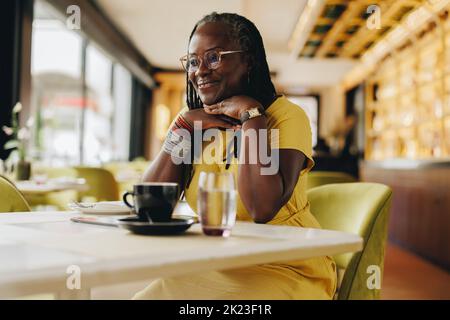 Glückliche reife Frau, die mit einem Lächeln wegschaut, während sie in einem Café sitzt. Fröhliche Frau mit Dreadlocks, die einige Zeit allein in einem Restaurant verbringt. Stockfoto