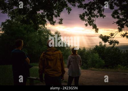 Drei Menschen beobachten den Sonnenuntergang, der in Wharfedale, Otley Chevin, West Yorkshire, England, grunzende Strahlen erzeugt Stockfoto