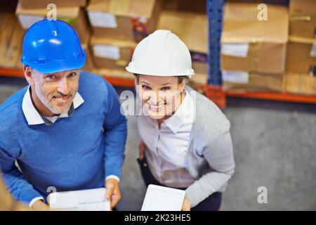 Wir haben das Zeug dazu. Aufnahme von zwei Personen in einem Lagerhaus, die Hardhüte trugen und die Kamera anlächelten. Stockfoto