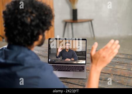 Nahaufnahme eines pakistanischen Mannes mit lockigen Haaren, der eine App für die Videokommunikation über die Distanz mit einem Kollegen, einem Freund, einem Online-Meeting und einem Laptop-Desktop mit männlichem Profil verwendet Stockfoto