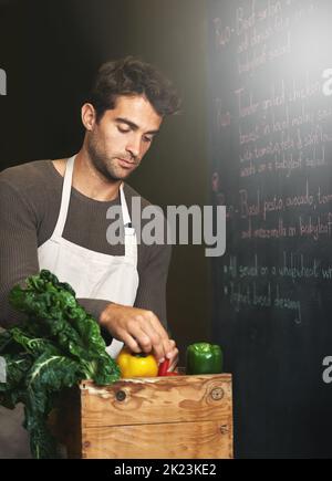 Er kauft nur Bio. Ein junger Mann, der in einer Küche mit einer Kiste voller frischem Gemüse vor ihm steht. Stockfoto