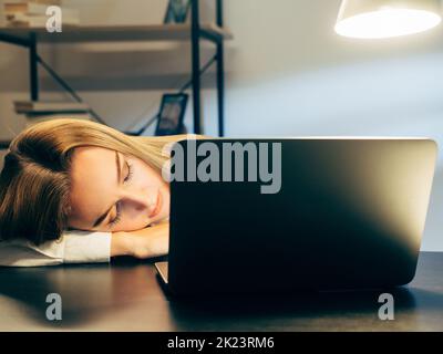 Burnout für die Arbeit. Ungesunde Arbeitslast. Freiberufler ermüdender Lebensstil. Erschöpfter Mitarbeiter schläft auf dem Schreibtisch vor dem Laptop im dunklen Arbeitsbereich des Büros. Stockfoto