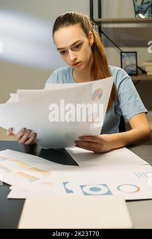 Überprüfung der Verantwortlichkeit. Freiberuflicher Mitarbeiter. Arbeiten von zu Hause aus. Fokussierte Frau, die spät in der Nacht im Büroarbeitsplatz sitzt und Papiere mit Grafiken studiert Stockfoto