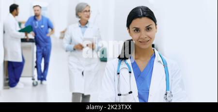 Asiatische Ärztin mit Stethoskop, das vor der Krankenhauslobby mit Ärzten in weißen Mänteln und medizinischem Personal steht. Arbeitstag in der Medizin Stockfoto