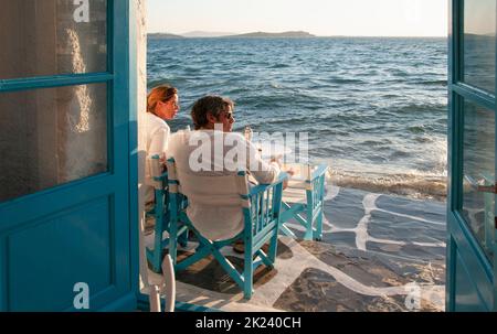 Ein junges Touristenpaar, das in einer Bar am Wasser auf Mykonos, Griechenland, sitzt und den Sonnenuntergang beobachtet Stockfoto