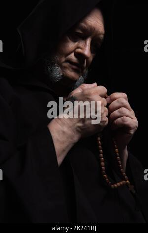 Senior tätowierten Mönch in Kapuze gehalten Rosenkranz während beten isoliert auf schwarzem, Stock Bild Stockfoto