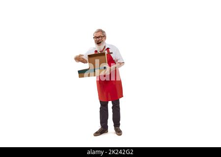 Porträt eines älteren Mannes, fröhlicher Verkäufer in roter Schürze, isoliert auf weißem Hintergrund. Geschenke verpacken Stockfoto